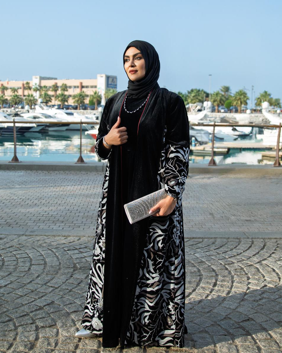 Woman in a black and white patterned abaya standing on a waterfront promenade.