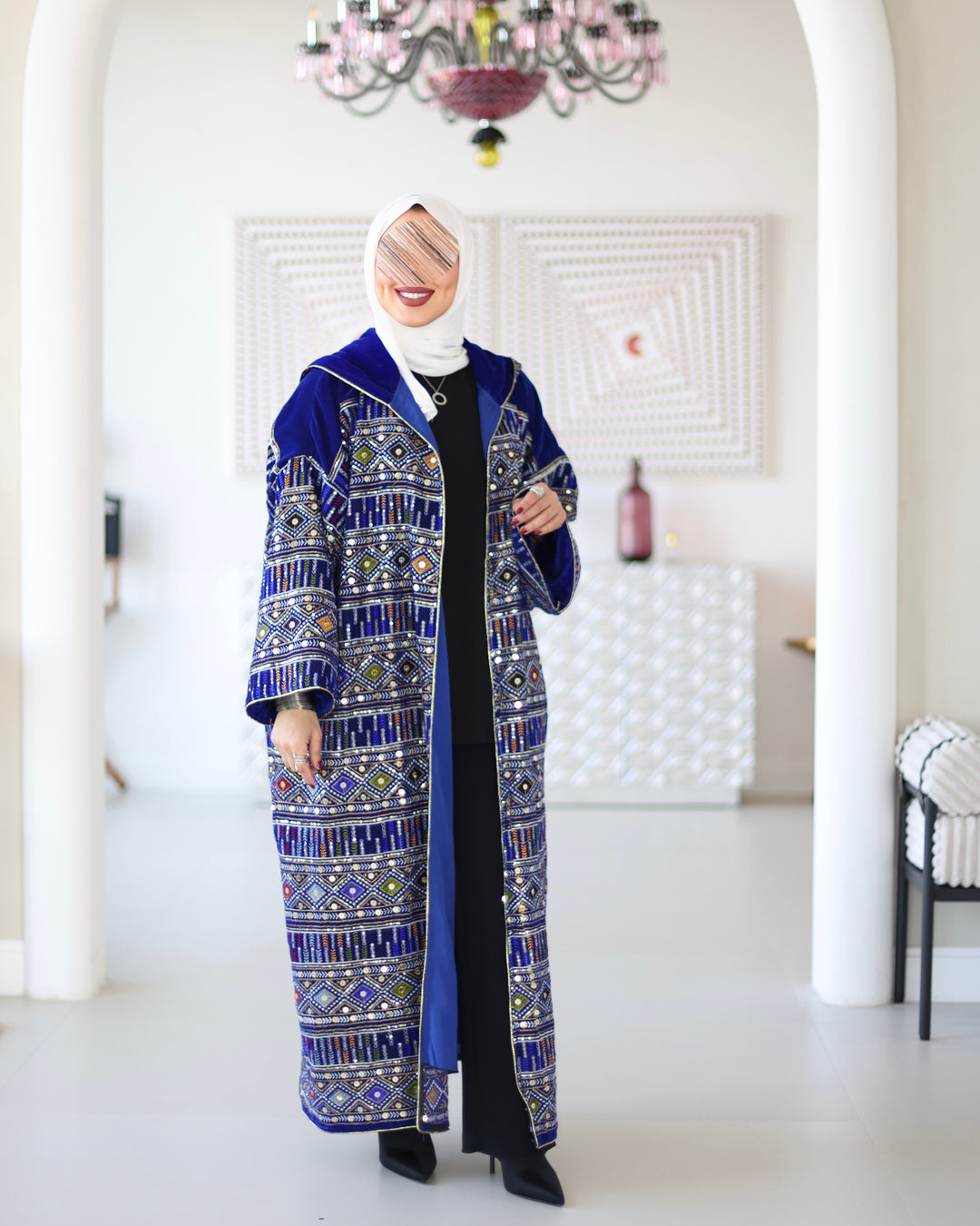 Woman wearing a long, patterned coat in a bright room with white walls and a chandelier.