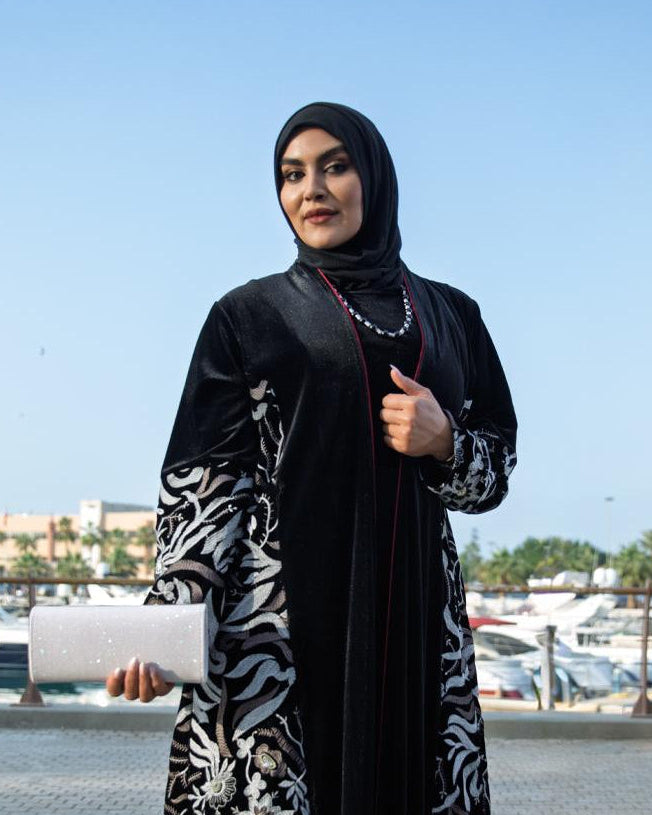 Woman in a black and white patterned abaya standing outdoors with a marina in the background.