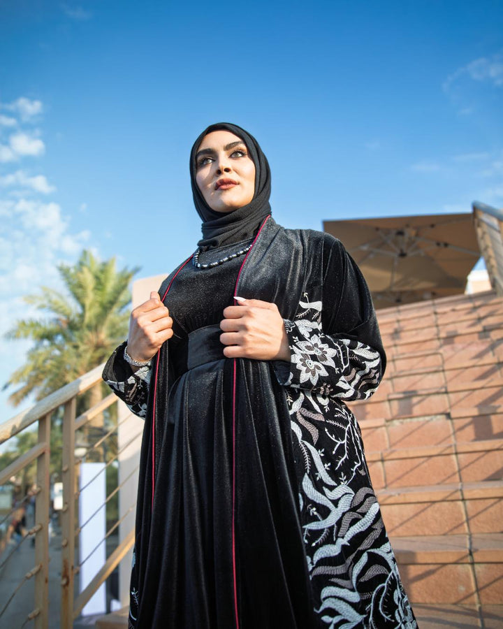 Woman wearing a black abaya with white patterns outdoors against a blue sky.