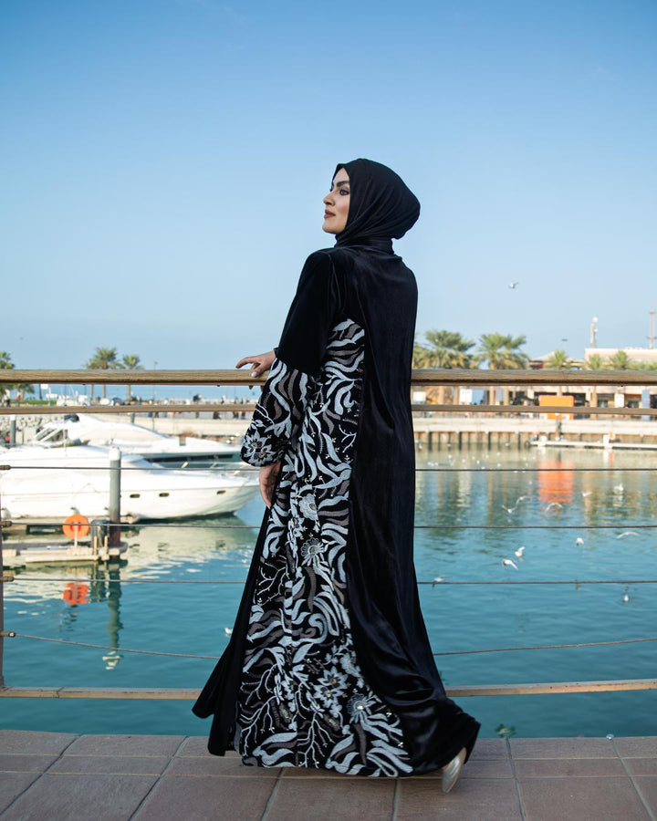 Woman in a black and white patterned abaya standing by a waterfront with boats and a clear sky.